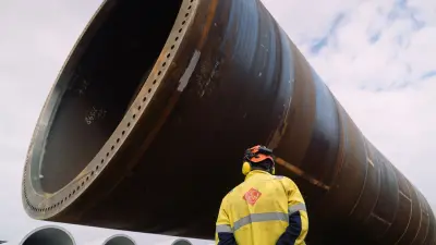 Ein Mitarbeiter im Hafen blickt von unten auf ein massives, liegendes Monopile-Fundament.