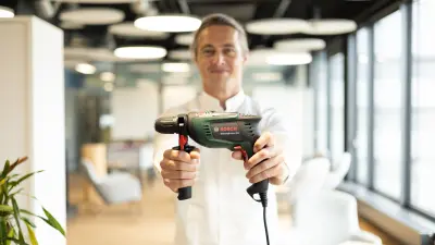 A smiling man holds a green Bosch UniversalImpact 800 impact drill directly towards the camera. In the blurry background, a modern office is visible.