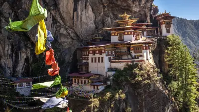 The Tiger's Nest Monastery (Paro Taktsang) in Bhutan, a stunning complex of white buildings with golden roofs, built into the side of a steep, rocky cliff face. Colorful prayer flags hang in the foreground on the left.